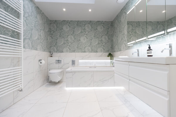 The images display a wallpaper with a large towel rail in white. A tiled floor and a vanity unit in white with a large mirror cabinet above it.